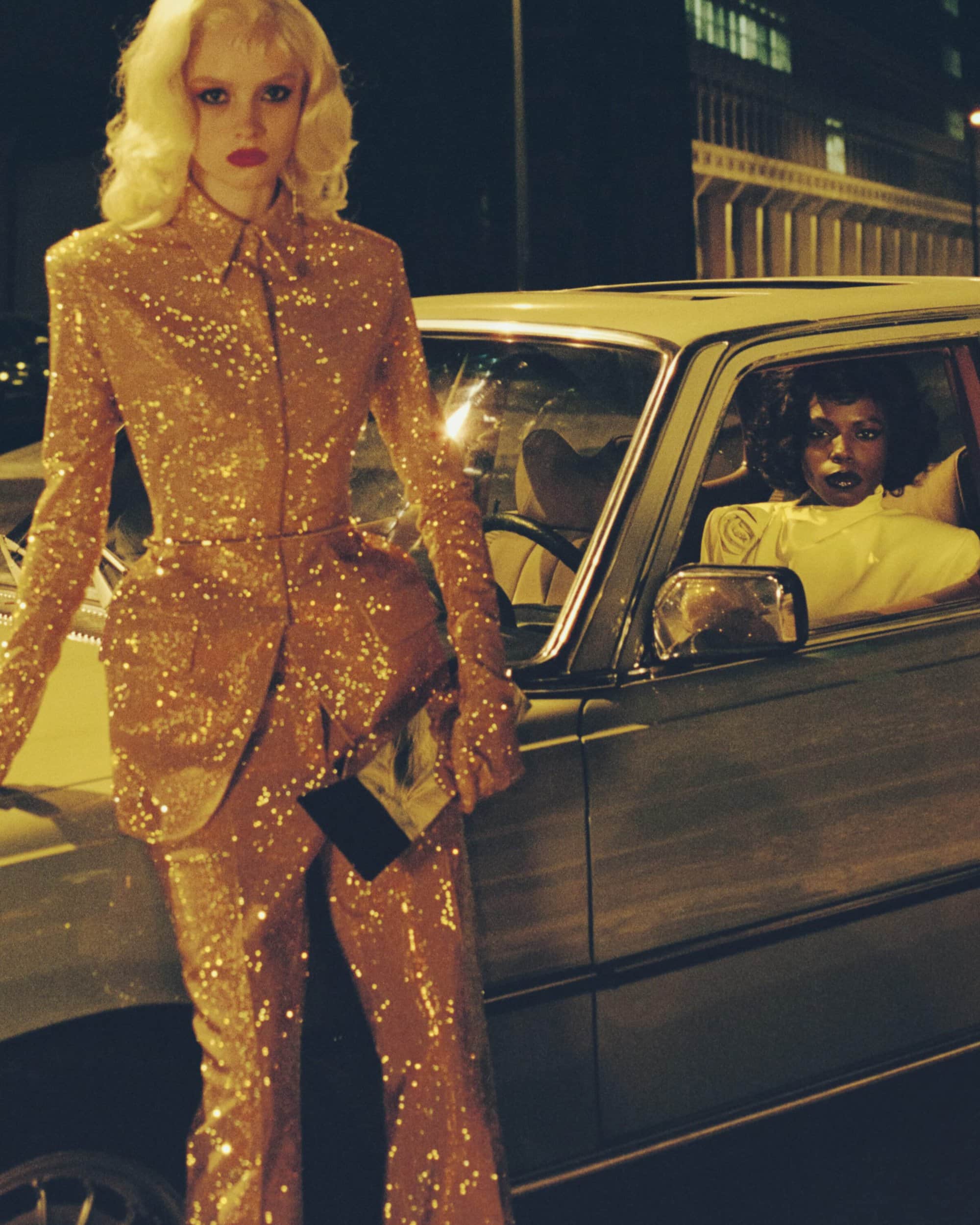 Two women pose near a vintage car at night: one in a gold sequin suit leaning on the car, the other sitting in the driver's seat.