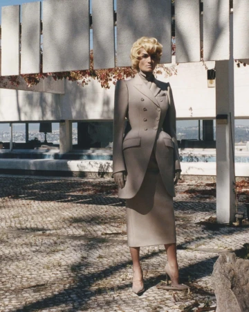 An adult female with blonde hair in a grey double-breasted suit with heels stands outside in front of a modern, geometric fountain.