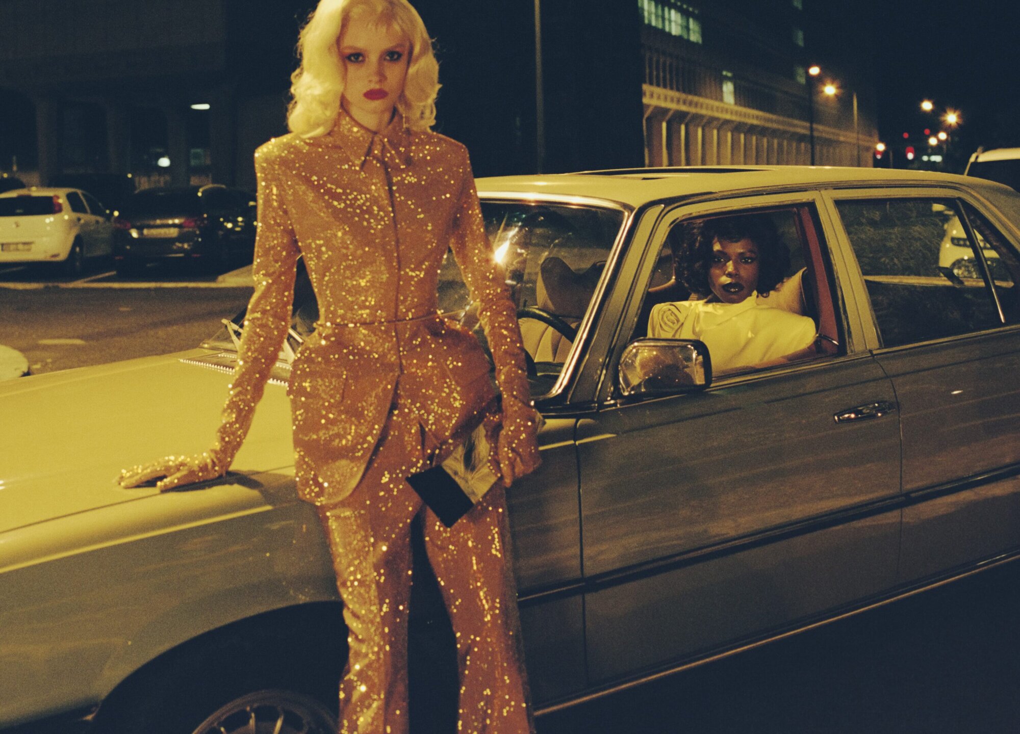 Two fashion models pose beside a classic car on a city street at night, one wearing a sparkling golden pantsuit, the other inside the vehicle.