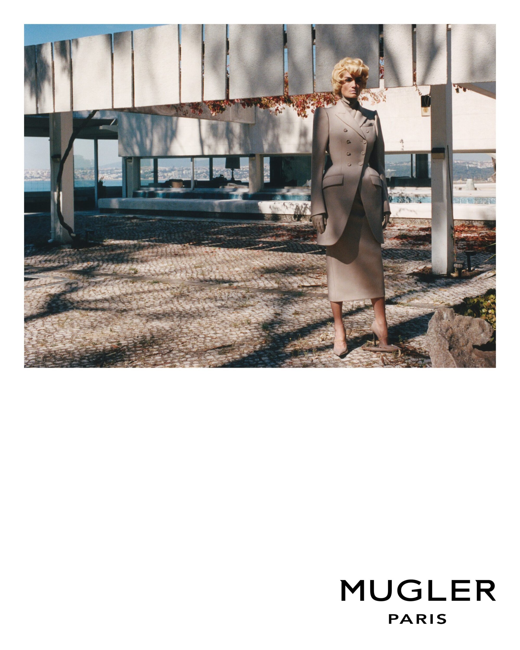 Woman in a tailored skirt suit posing near a modern concrete building with a glimpse of the ocean in the background during daytime.