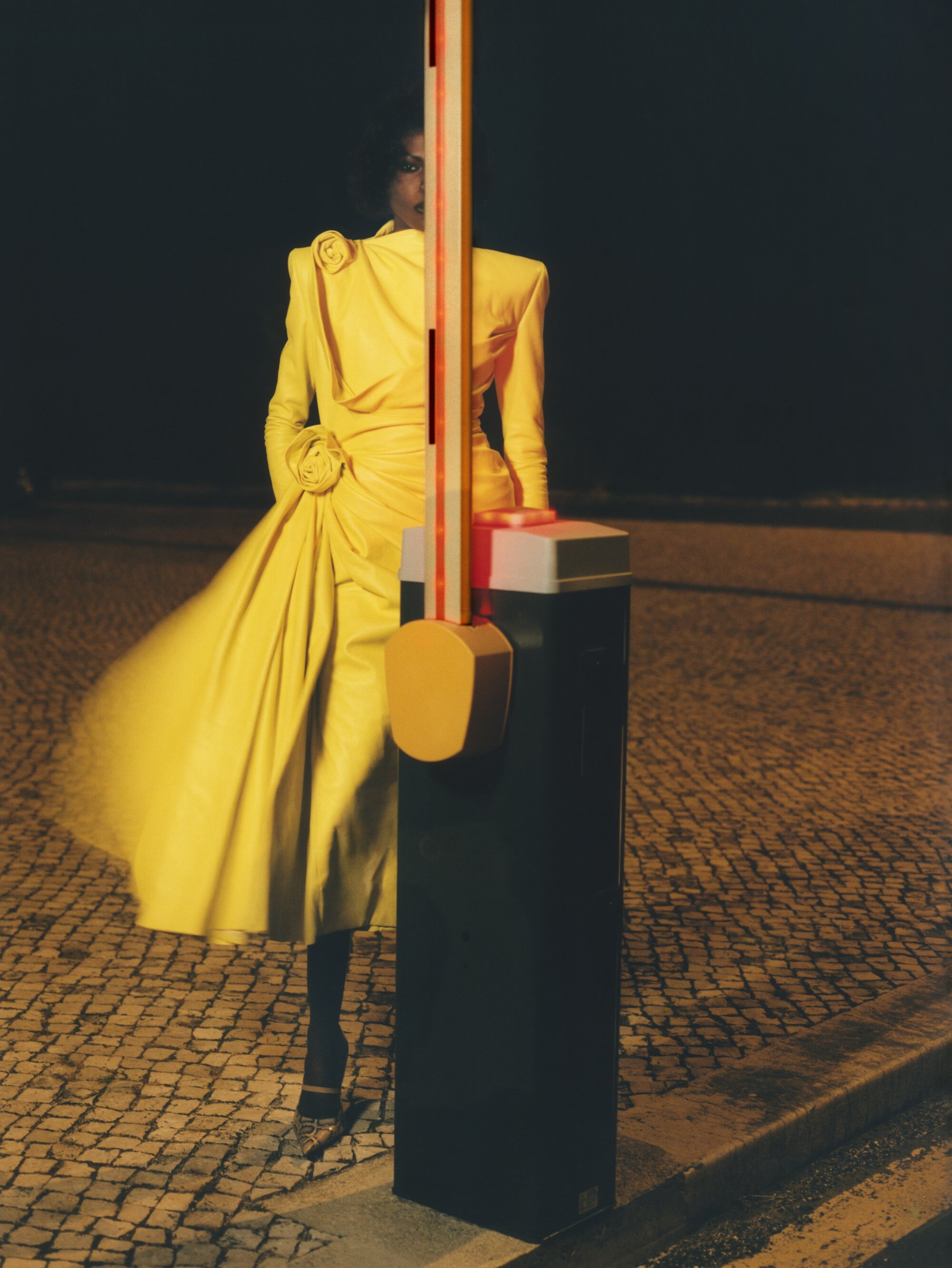 Model standing behind a vertical security barrier wearing a bright yellow dress at night