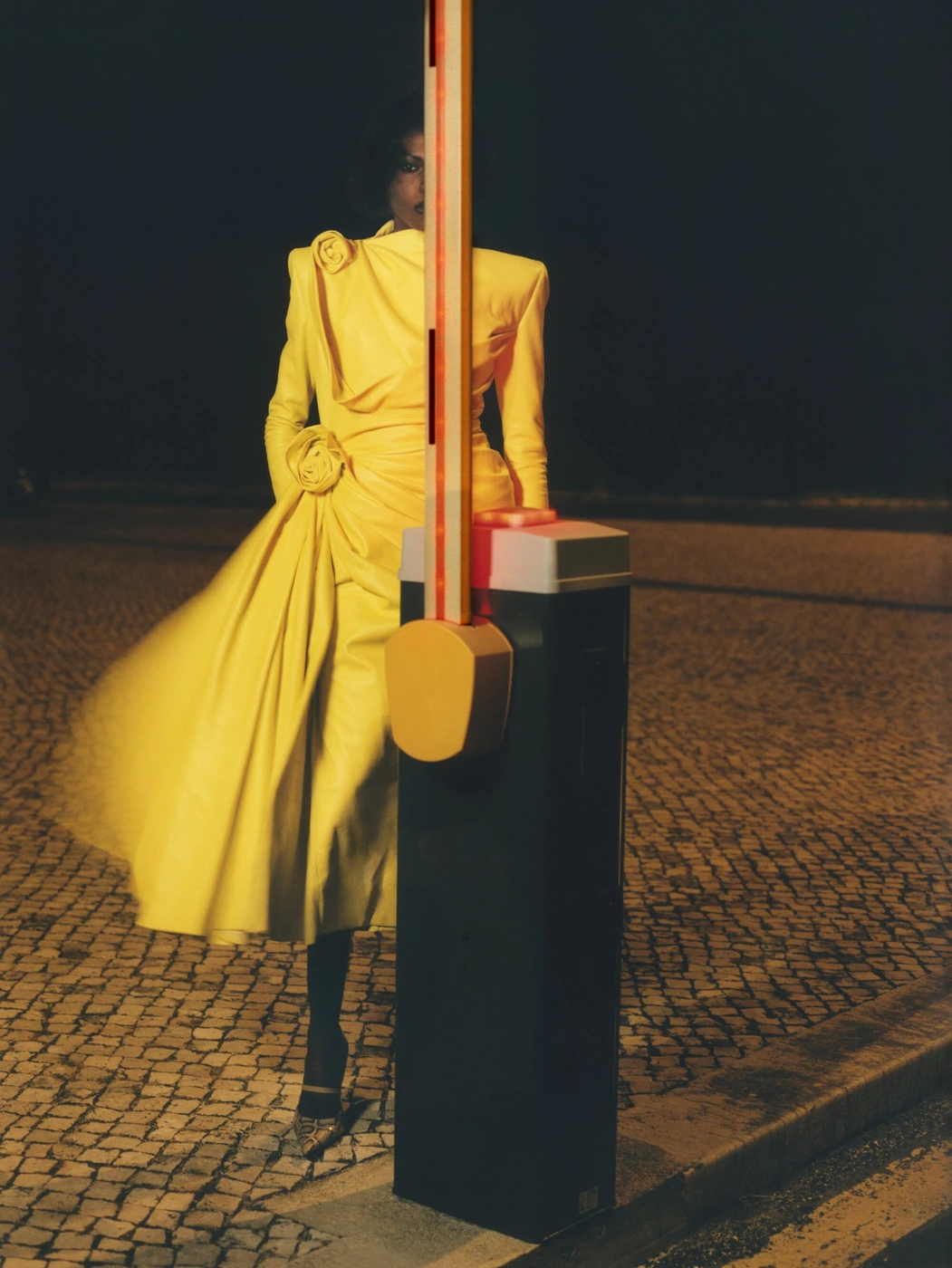 Model standing behind a vertical security barrier wearing a bright yellow dress at night