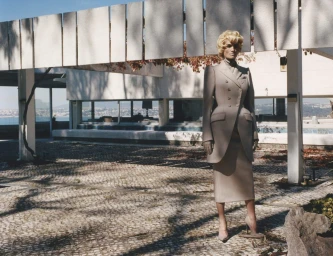 Model standing outdoors wearing a tailored beige skirt suit and heels beneath modern architectural structures