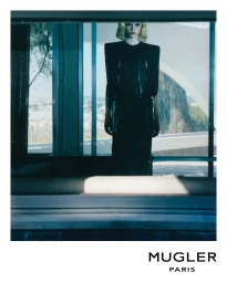 Model with blonde hair wearing a structured black Mugler dress standing inside near a glass window