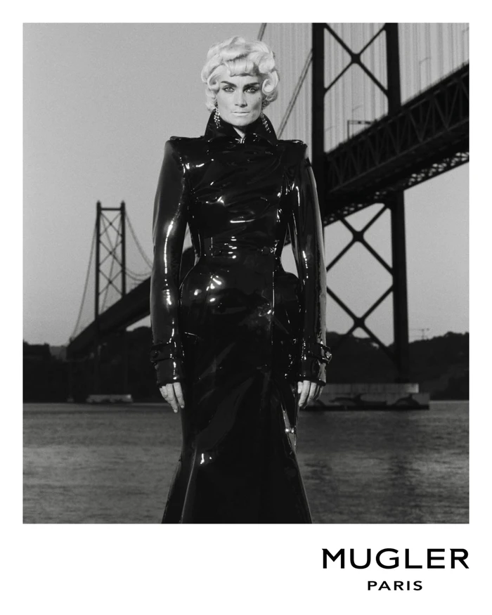 Monochrome fashion portrait of a model in a glossy black vinyl coat standing before a large suspension bridge