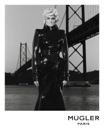 Monochrome fashion portrait of a model in a glossy black vinyl coat standing before a large suspension bridge