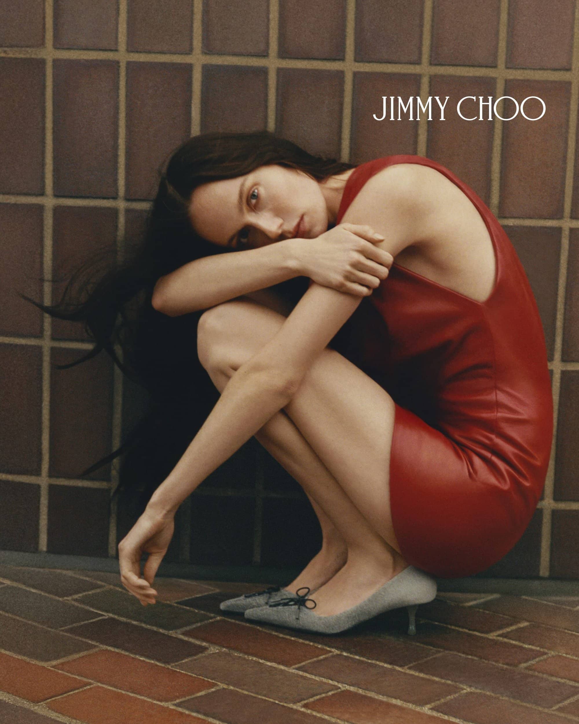 A woman in a red dress and grey kitten heels crouches against a brick wall in a fashion editorial for Jimmy Choo.