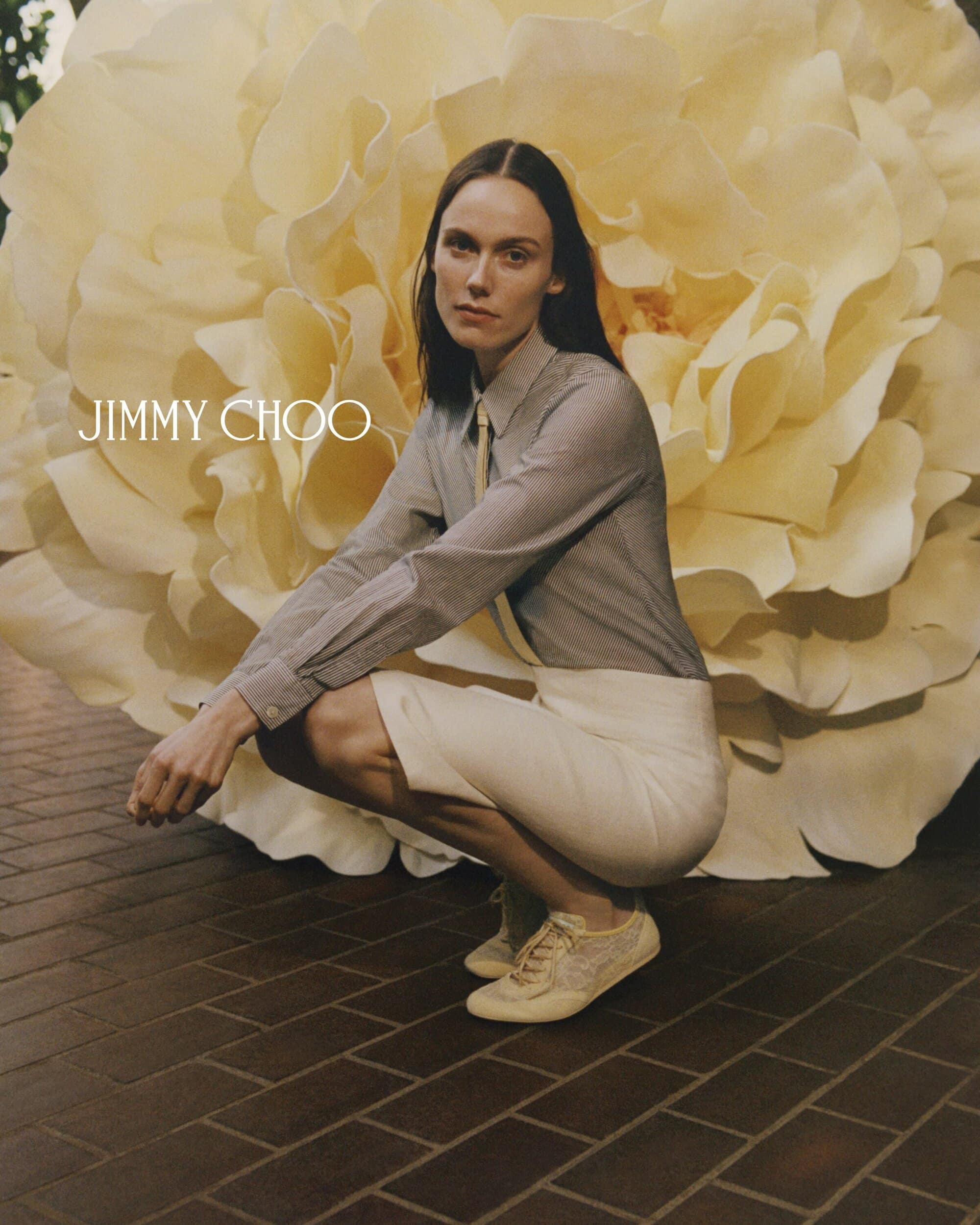 An adult female crouches in front of a massive faux flower while advertising Jimmy Choo sneakers and a business-casual look.