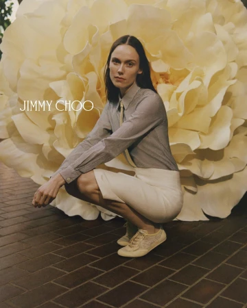 An adult female crouches in front of a massive faux flower while advertising Jimmy Choo sneakers and a business-casual look.
