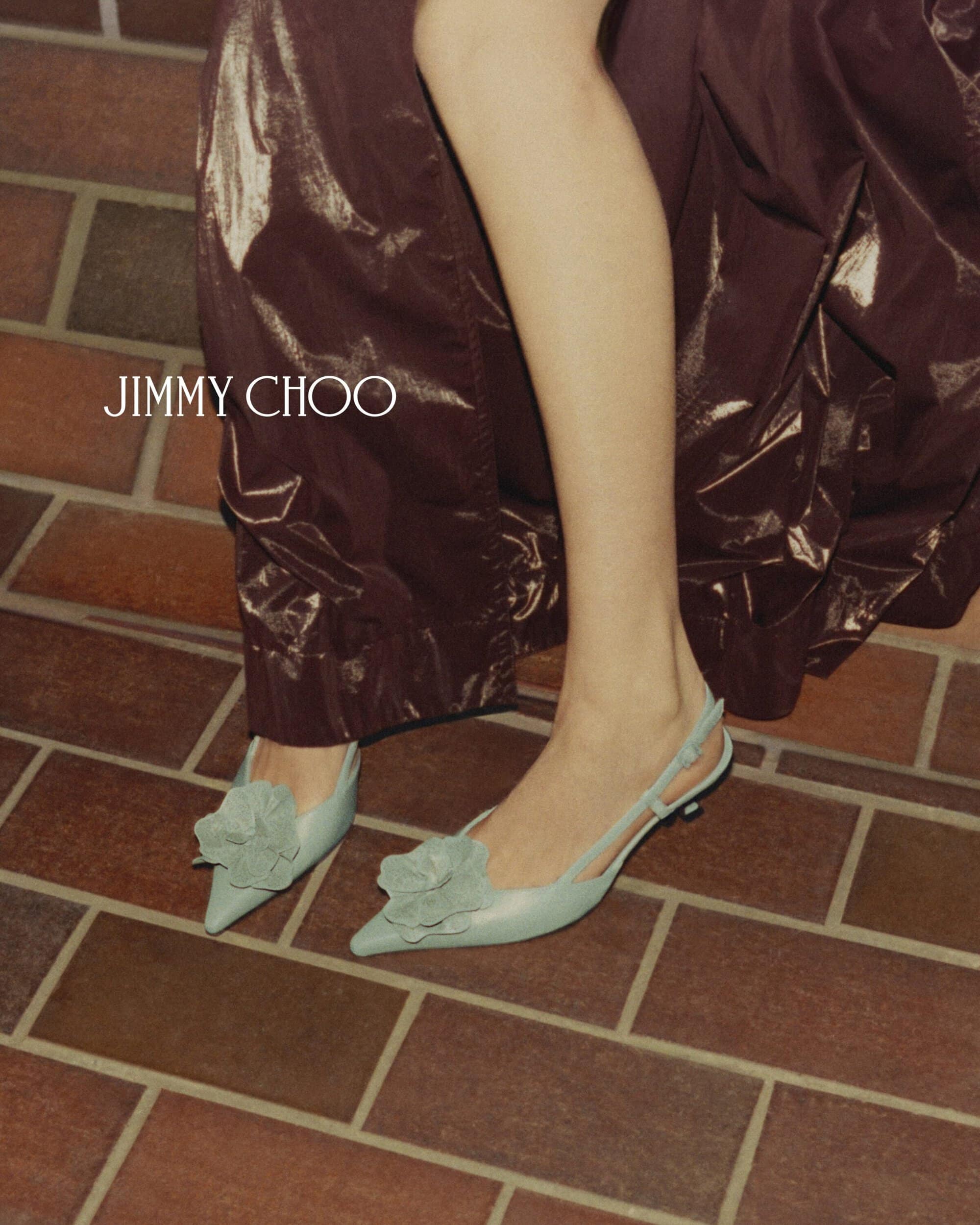 Close-up of a slender woman's legs and feet wearing light blue Jimmy Choo slingback heels with a floral detail. The shoes are on a brick surface.