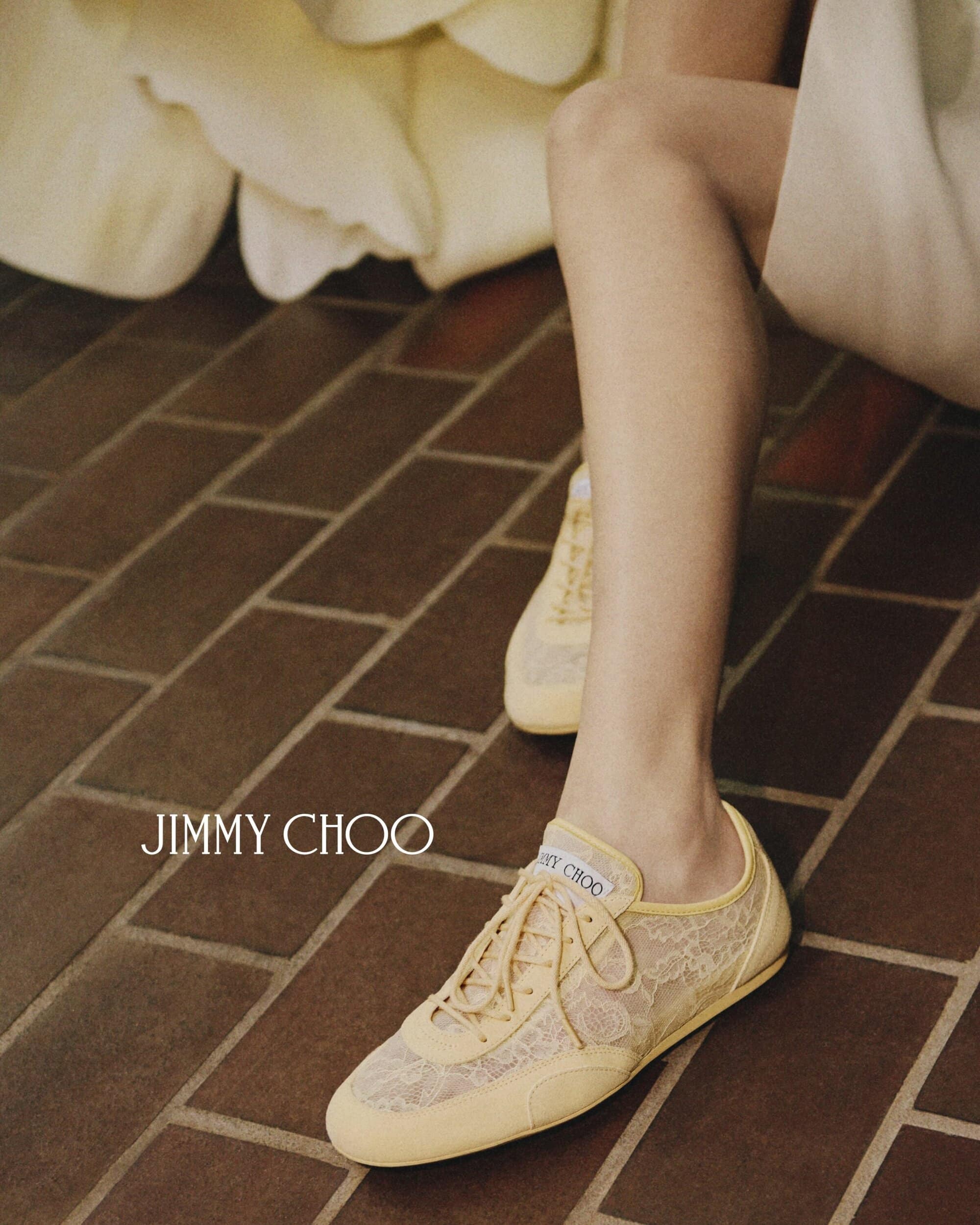 Close-up of Jimmy Choo lace sneakers on brick surface with a woman's legs visible and white flower detail in the background.