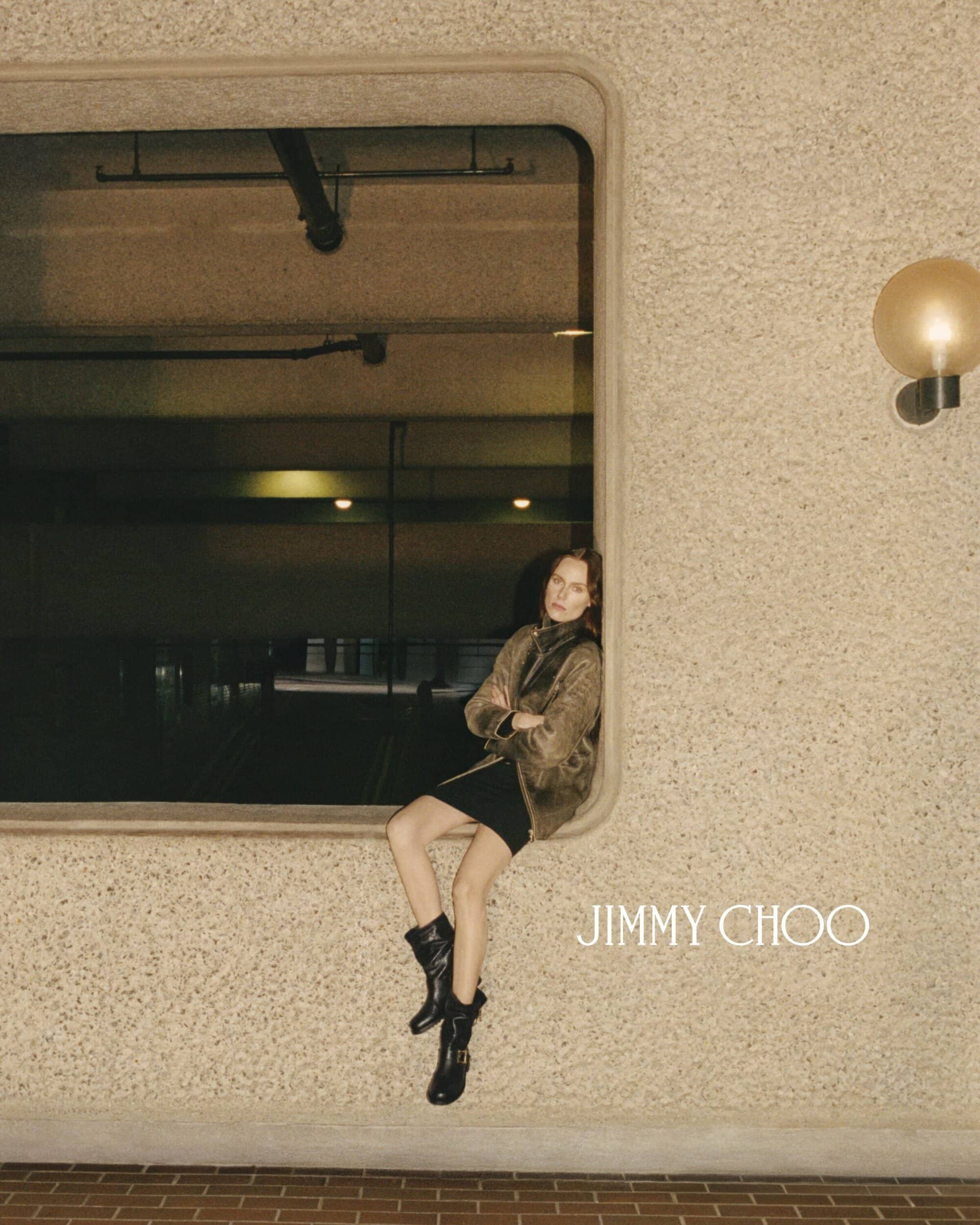 Fashion shoot of a woman sitting in a window wearing black boots and a jacket, with the Jimmy Choo logo on a textured wall.