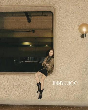 Fashion shoot of a woman sitting in a window wearing black boots and a jacket, with the Jimmy Choo logo on a textured wall.