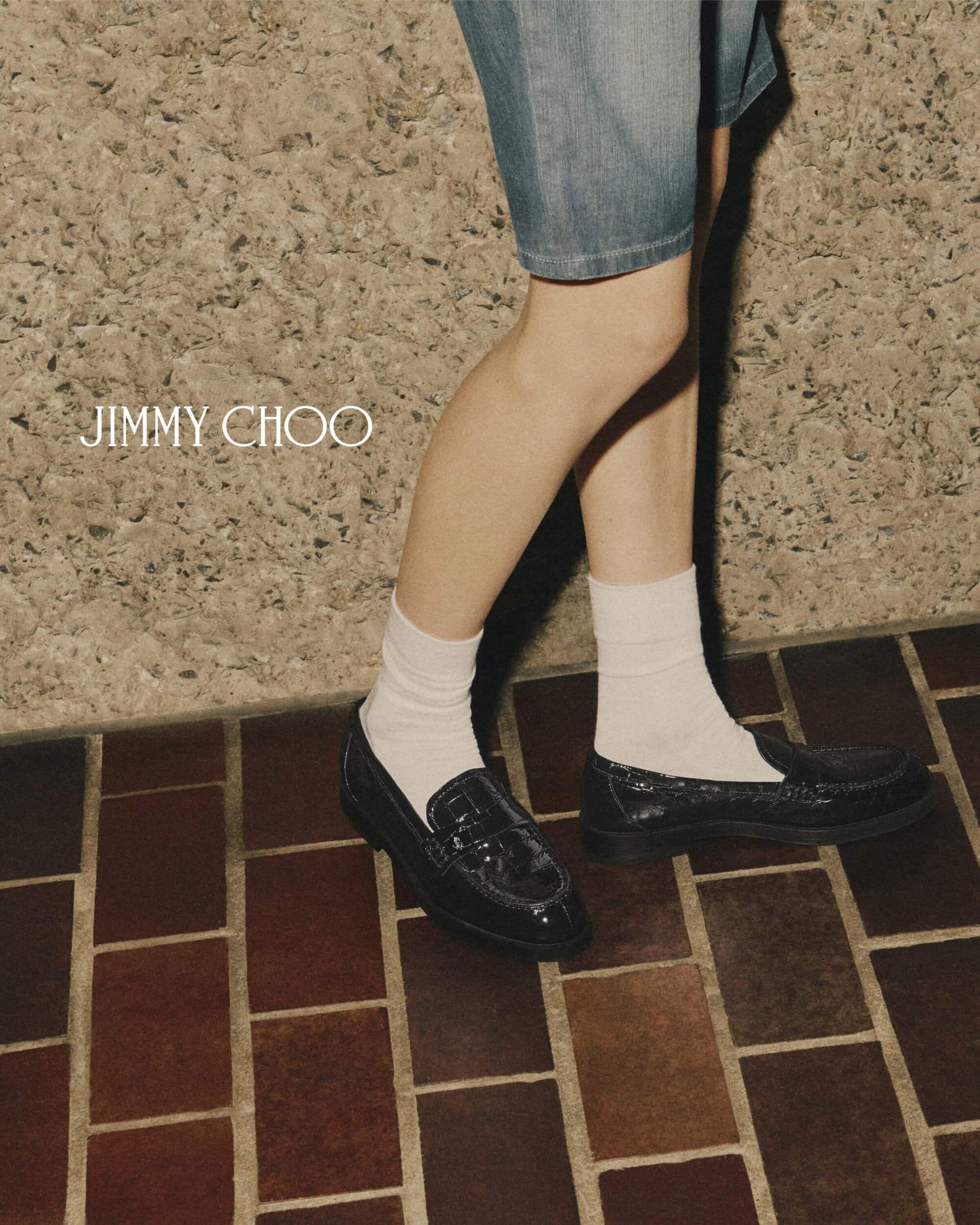 Fashion shot featuring a person wearing denim shorts, white socks, and black loafers by Jimmy Choo, standing on a brick surface.