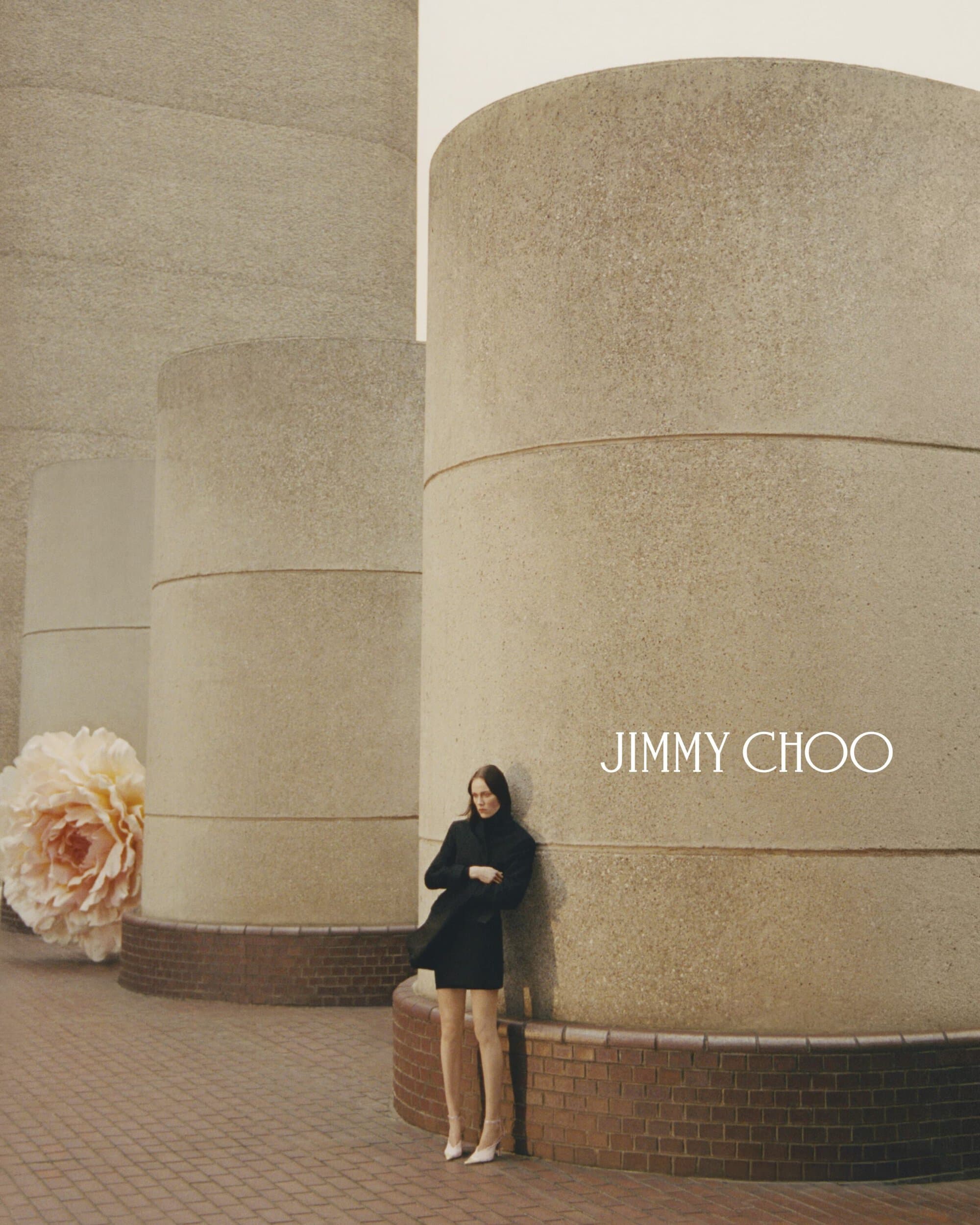 Woman in black coat and nude hosiery poses by cylindrical concrete columns with a large beige flower and the Jimmy Choo logo.