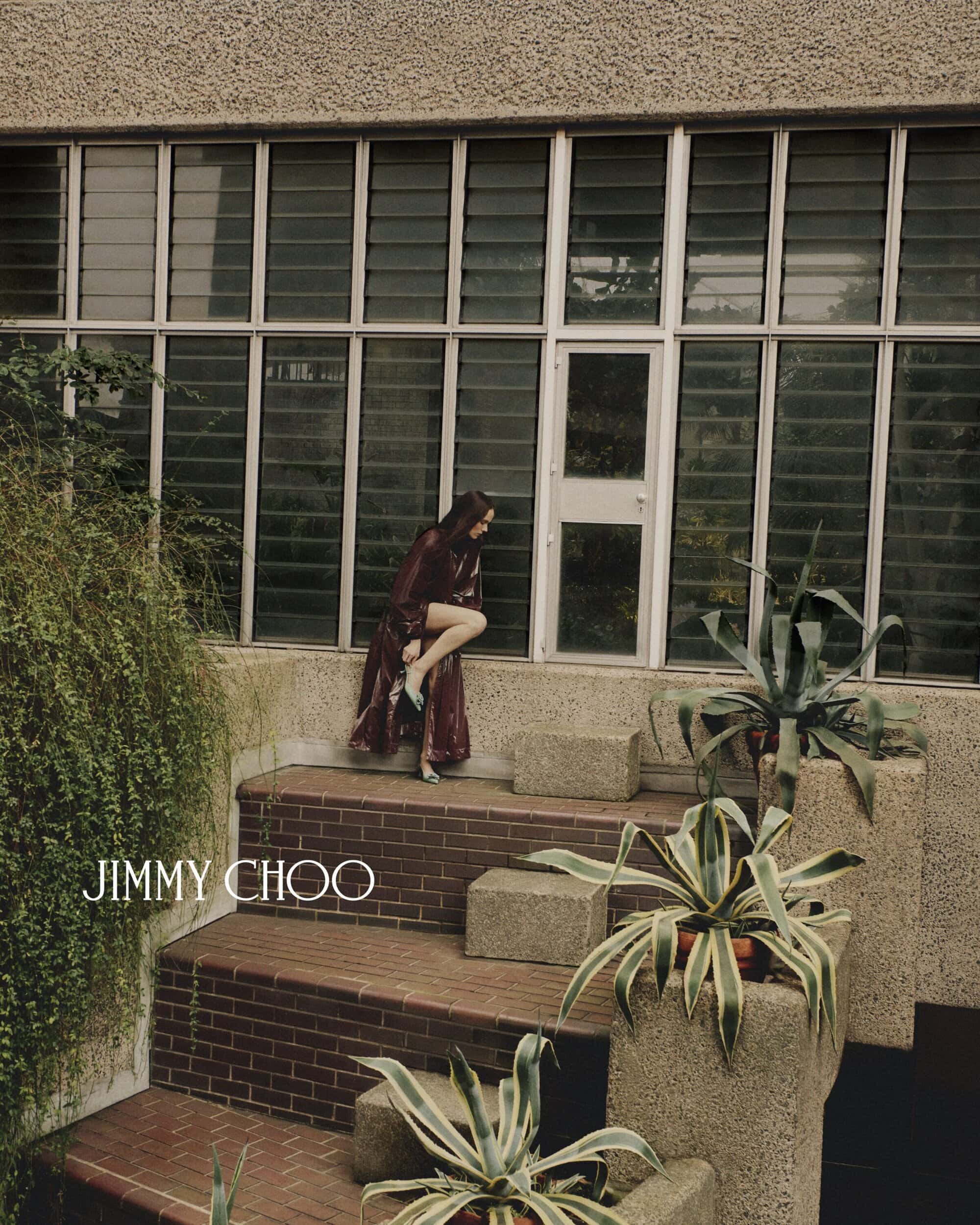 Woman in a maroon dress and light blue shoes poses on steps of a building with plants, with the Jimmy Choo logo.