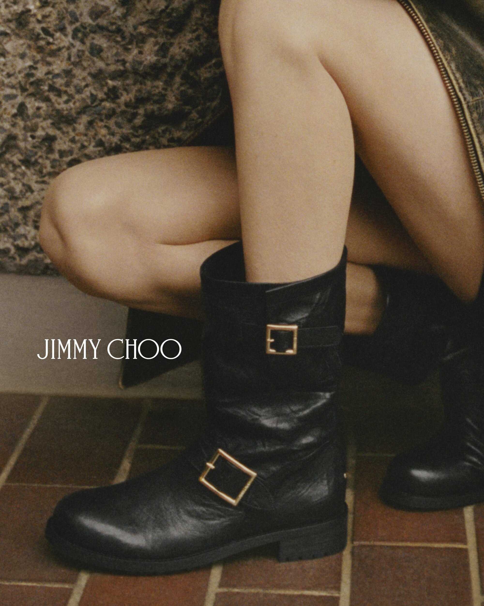 A person wearing black leather biker boots with gold buckles from Jimmy Choo. The image is taken indoors on a tiled surface.
