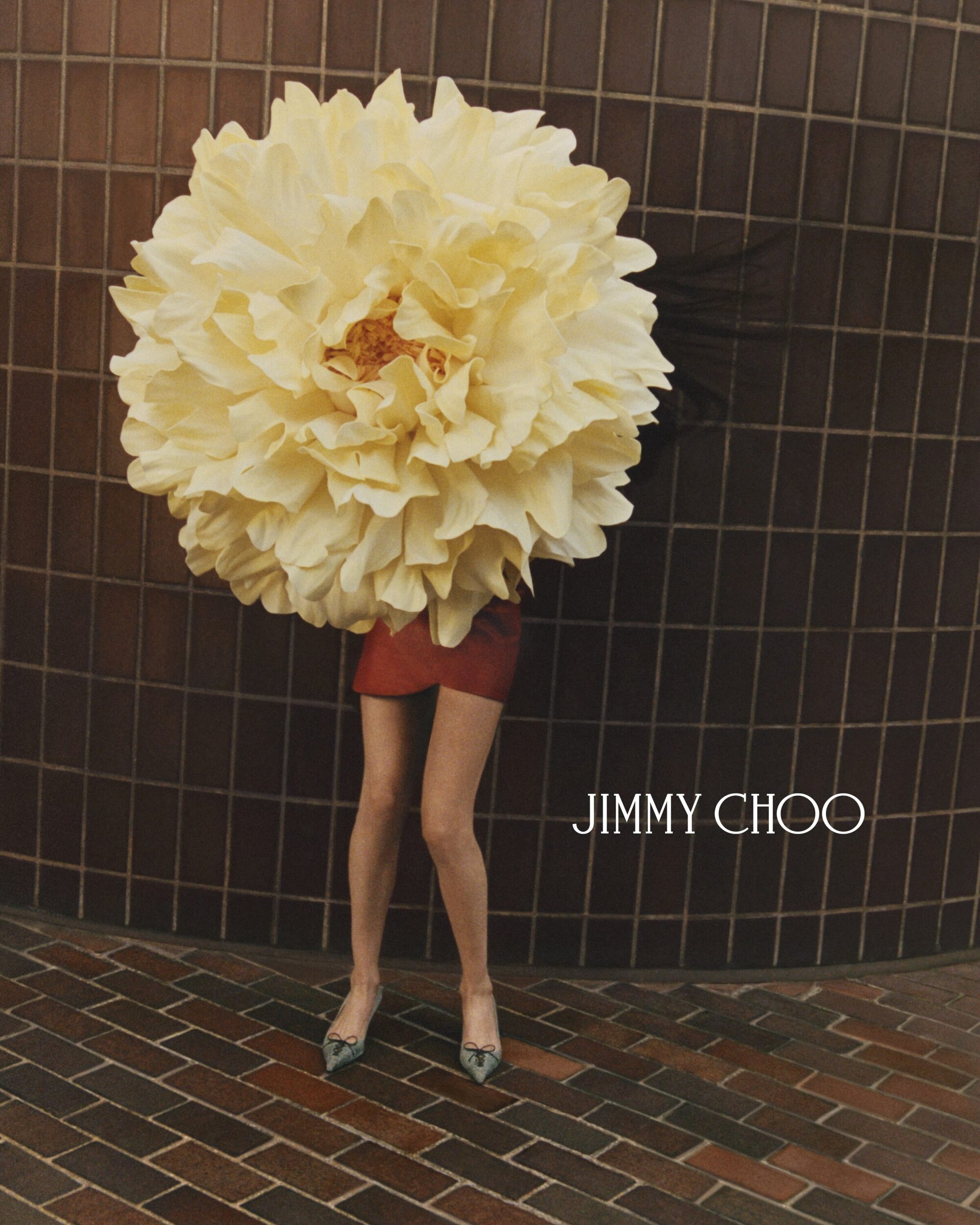 Model wearing a giant pale yellow flower headpiece, red skirt, and heels against a dark tiled wall for Jimmy Choo.
