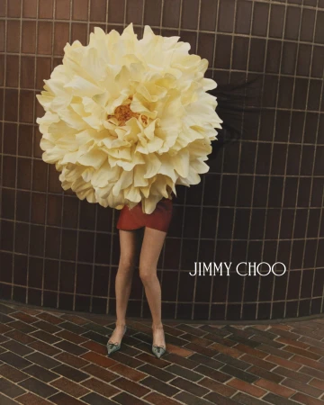 Model wearing a giant pale yellow flower headpiece, red skirt, and heels against a dark tiled wall for Jimmy Choo.
