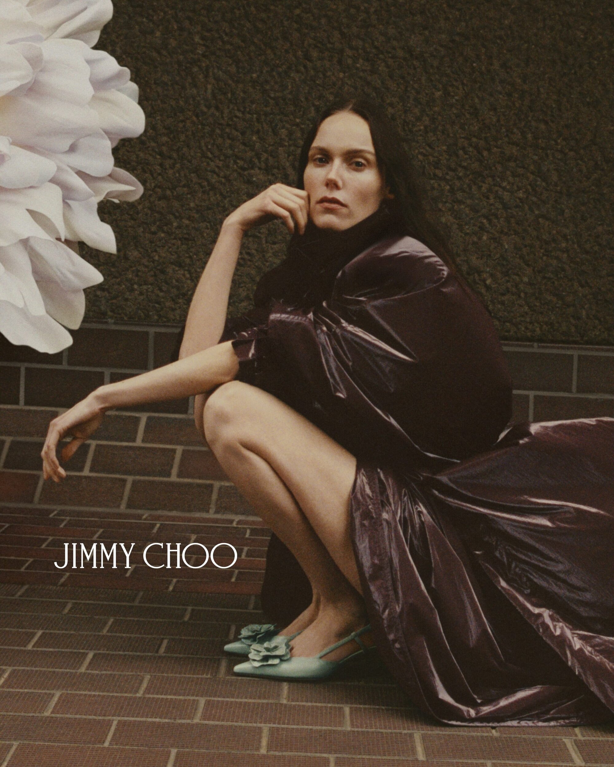 Fashion ad: Woman in sculptural dress poses with Jimmy Choo heels on brick. Large white floral element on dark green background.