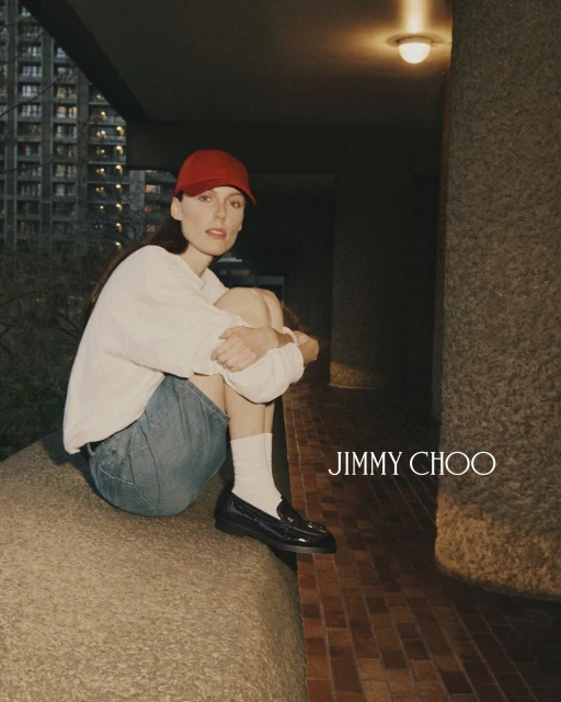 A woman in a red cap and light-colored sweatshirt sits on a concrete ledge with her legs tucked up; Jimmy Choo logo to the right.