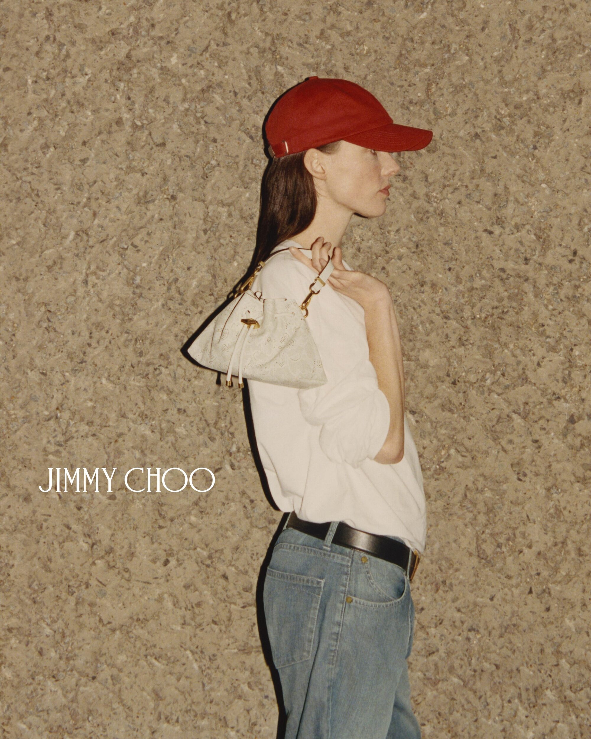 Fashion shot of a woman wearing a red baseball cap, light blue jeans, and a white top, holding a small purse in an indoor setting.