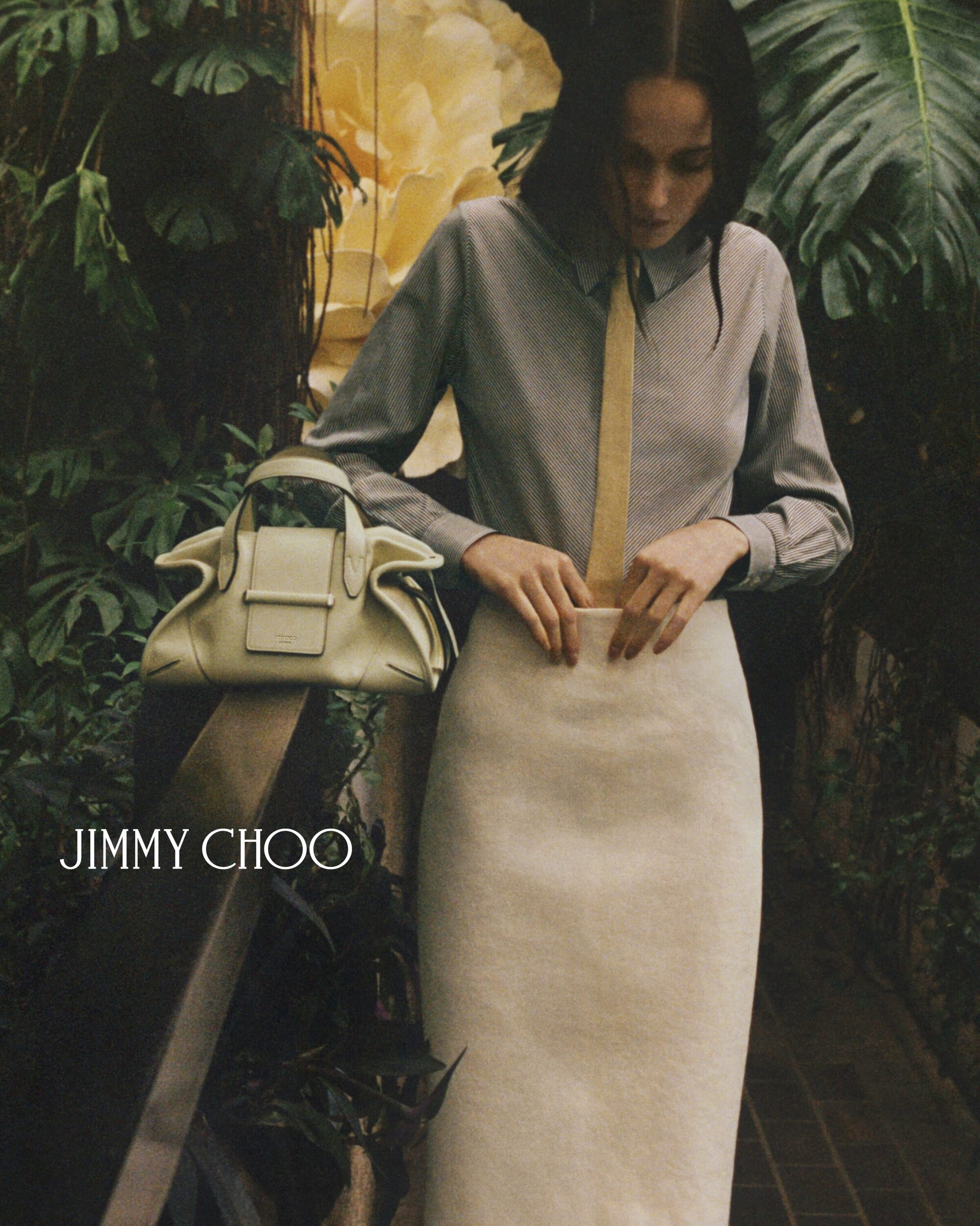Woman wearing a gray striped shirt, beige skirt, and tan tie, posing with a cream-colored Jimmy Choo purse in a botanical garden setting.