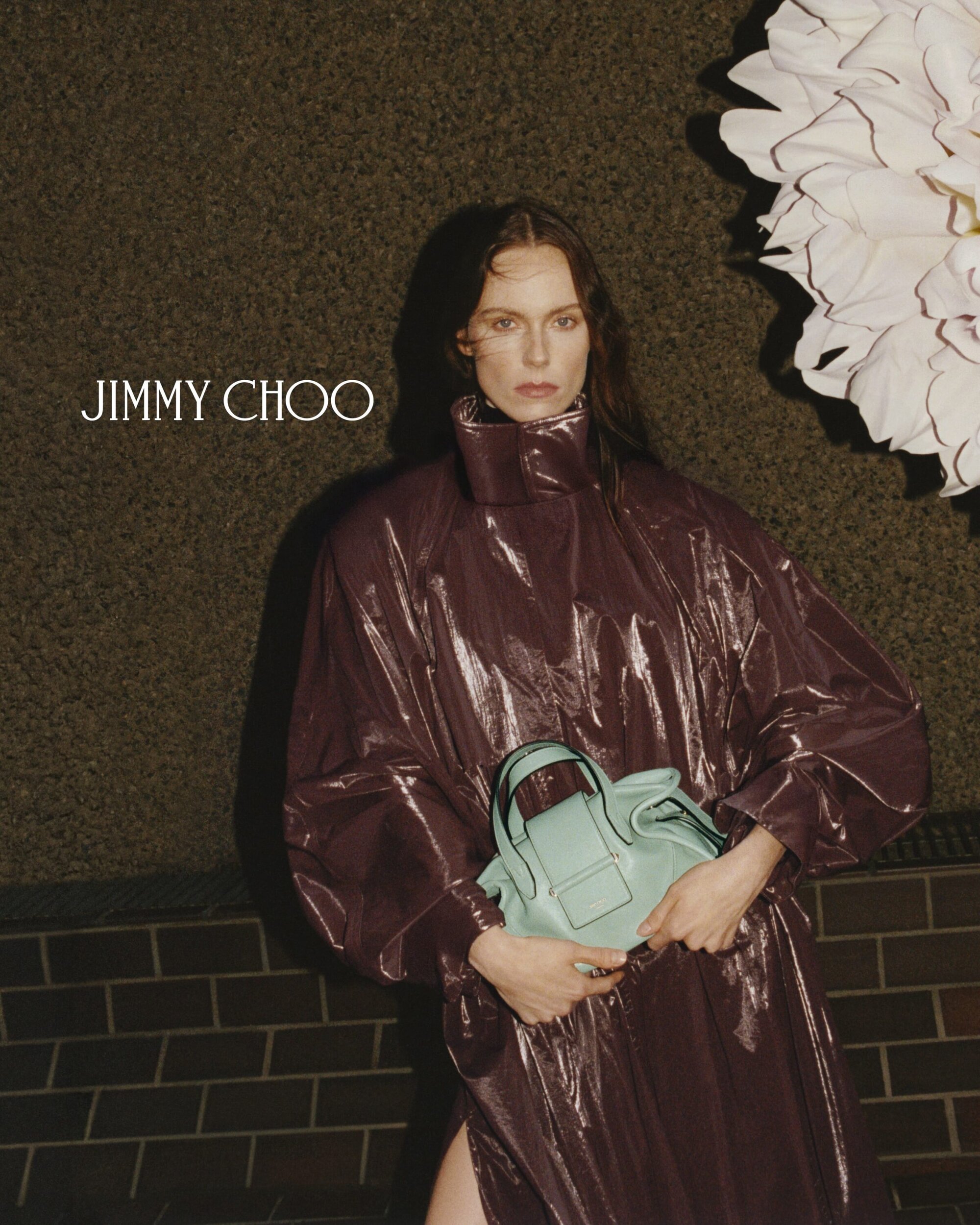 Fashion editorial shot of a woman in a glossy burgundy coat holding a light blue purse in front of a textured green wall and flower.