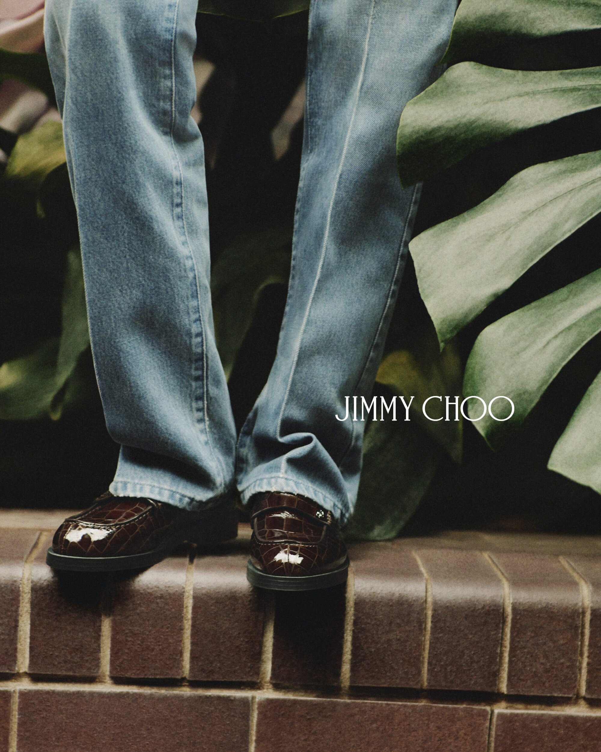 A stylish composition features a person's legs in jeans, showcasing brown leather loafers against a backdrop of lush green leaves.