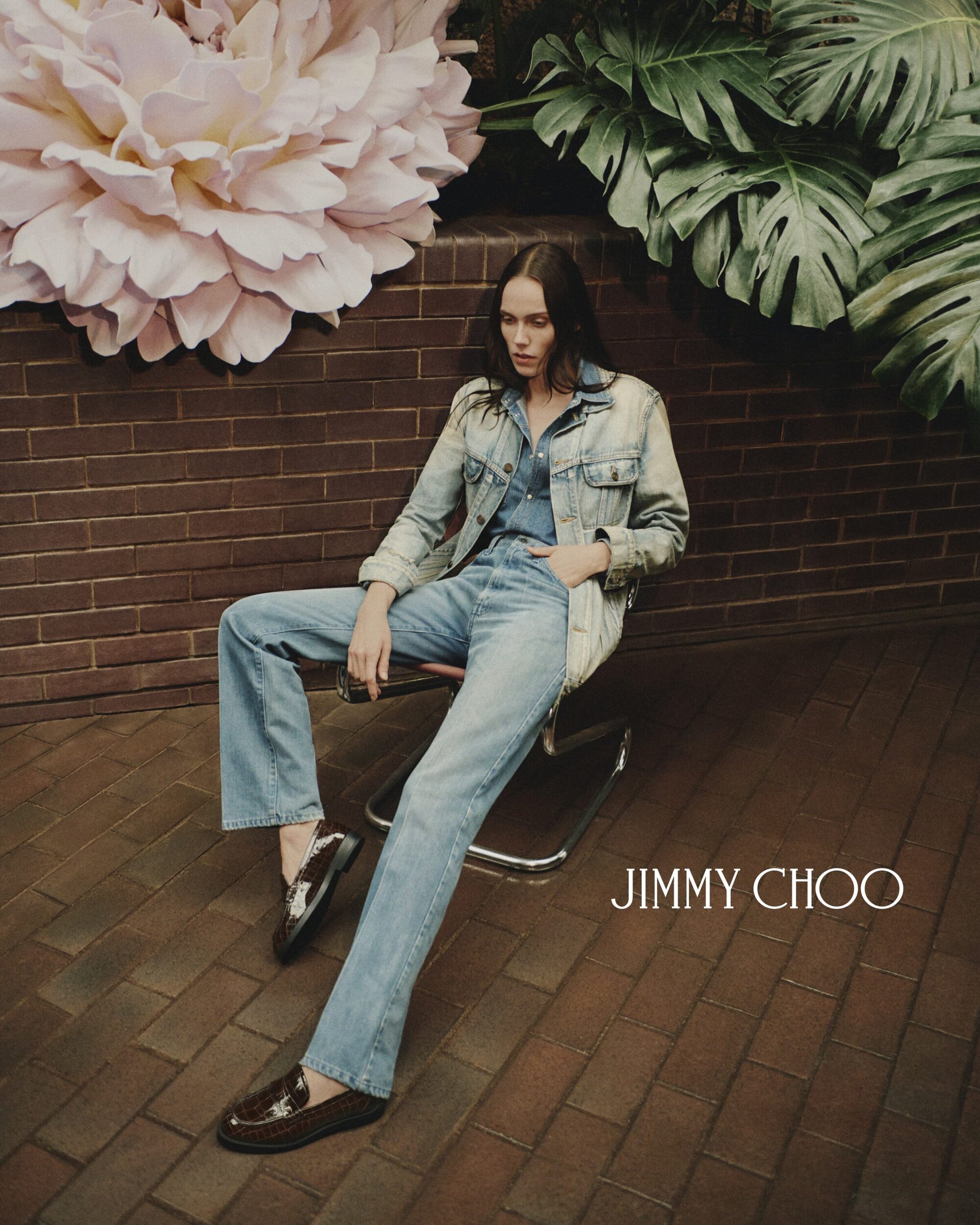 A young woman poses in a denim outfit, seated in front of a brick wall with a floral and monstera-leaf background for a Jimmy Choo ad.