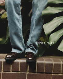 Close-up of blue jeans and brown crocodile-embossed loafers standing on a ledge next to large tropical leaves.
