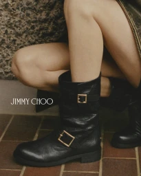 Close-up of black leather boots with gold buckles worn by a model against a textured tiled floor and textured wall