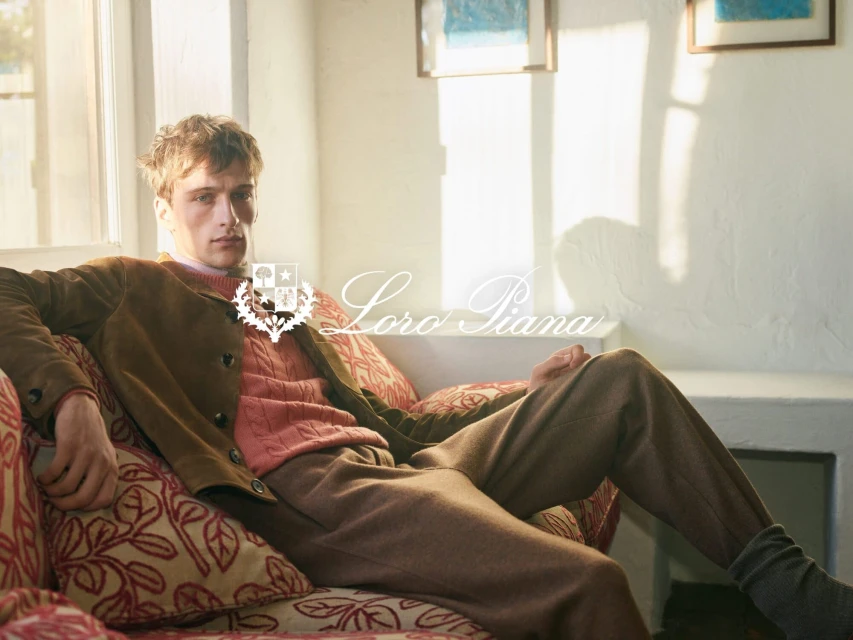 A blonde man in a brown jacket and pants sits on a patterned couch in a well-lit room, showcasing an elegant and comfortable fashion style.