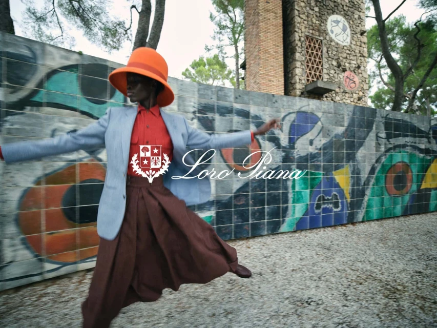 Fashionable Black woman in an orange hat, grey blazer, and brown skirt poses dynamically in front of a mural-covered wall and tower.