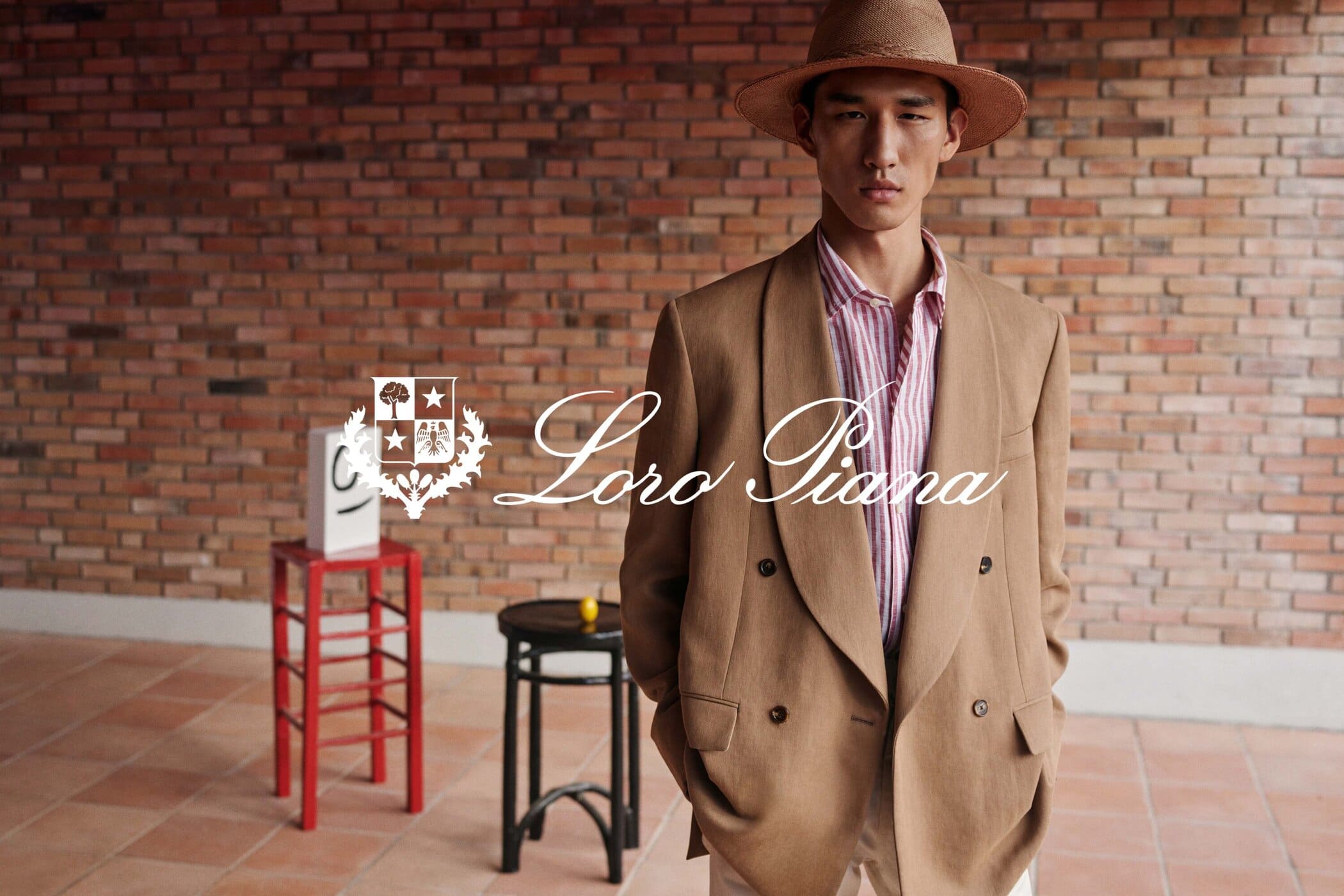 Asian male model wears a straw hat and brown double-breasted blazer in a fashion editorial with a brick wall backdrop and logo.