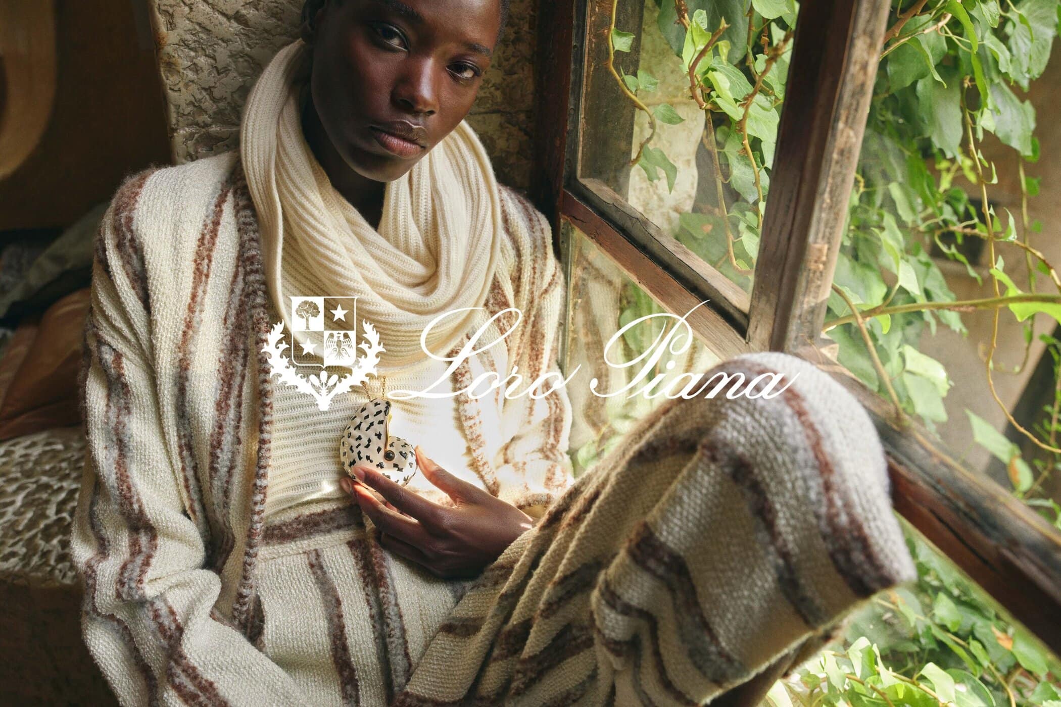 A dark-skinned woman poses in a beige-striped knit outfit by a window with climbing plants, holding a decorative object and glancing at the viewer.