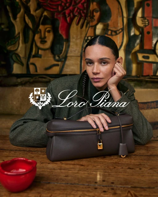 Brunette woman in a green tweed jacket with a brown leather handbag on a wooden table in front of an abstract painting.