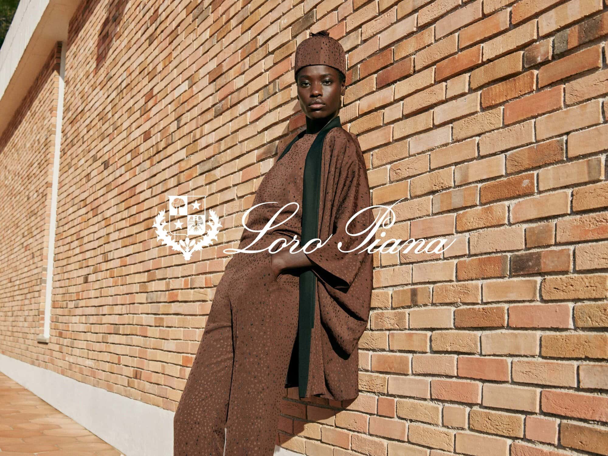A woman wearing a brown outfit and hat poses against a brick wall. The fashion brand Loro Piana logo is in the foreground.