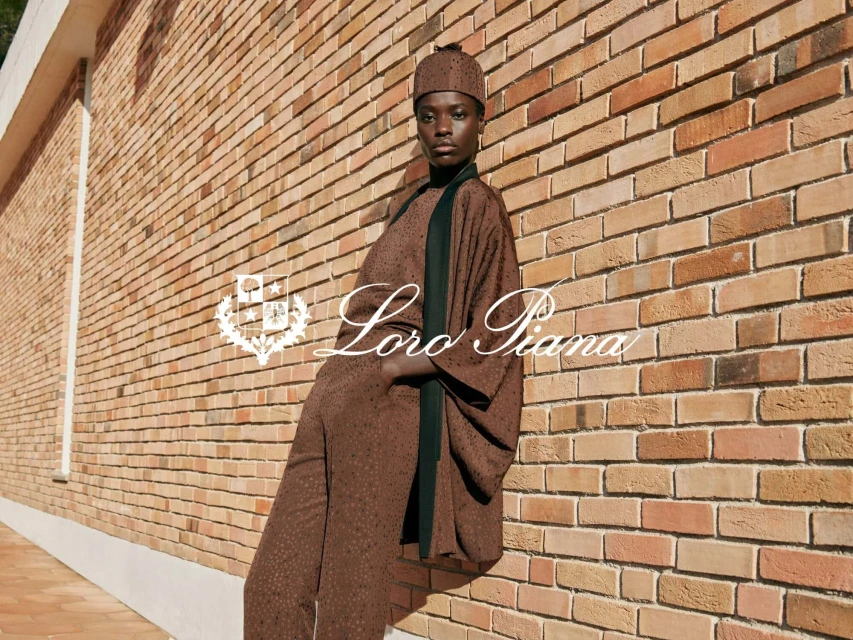 A woman wearing a brown outfit and hat poses against a brick wall. The fashion brand Loro Piana logo is in the foreground.