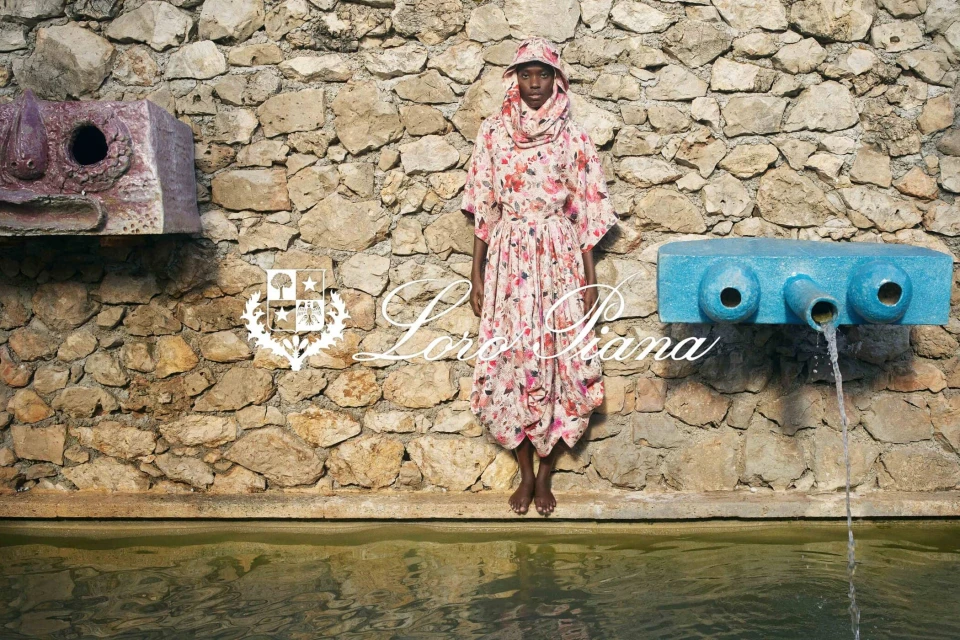 Fashion shot of an adult woman wearing a floral patterned dress, headscarf and standing in front of a stone wall with water feature.