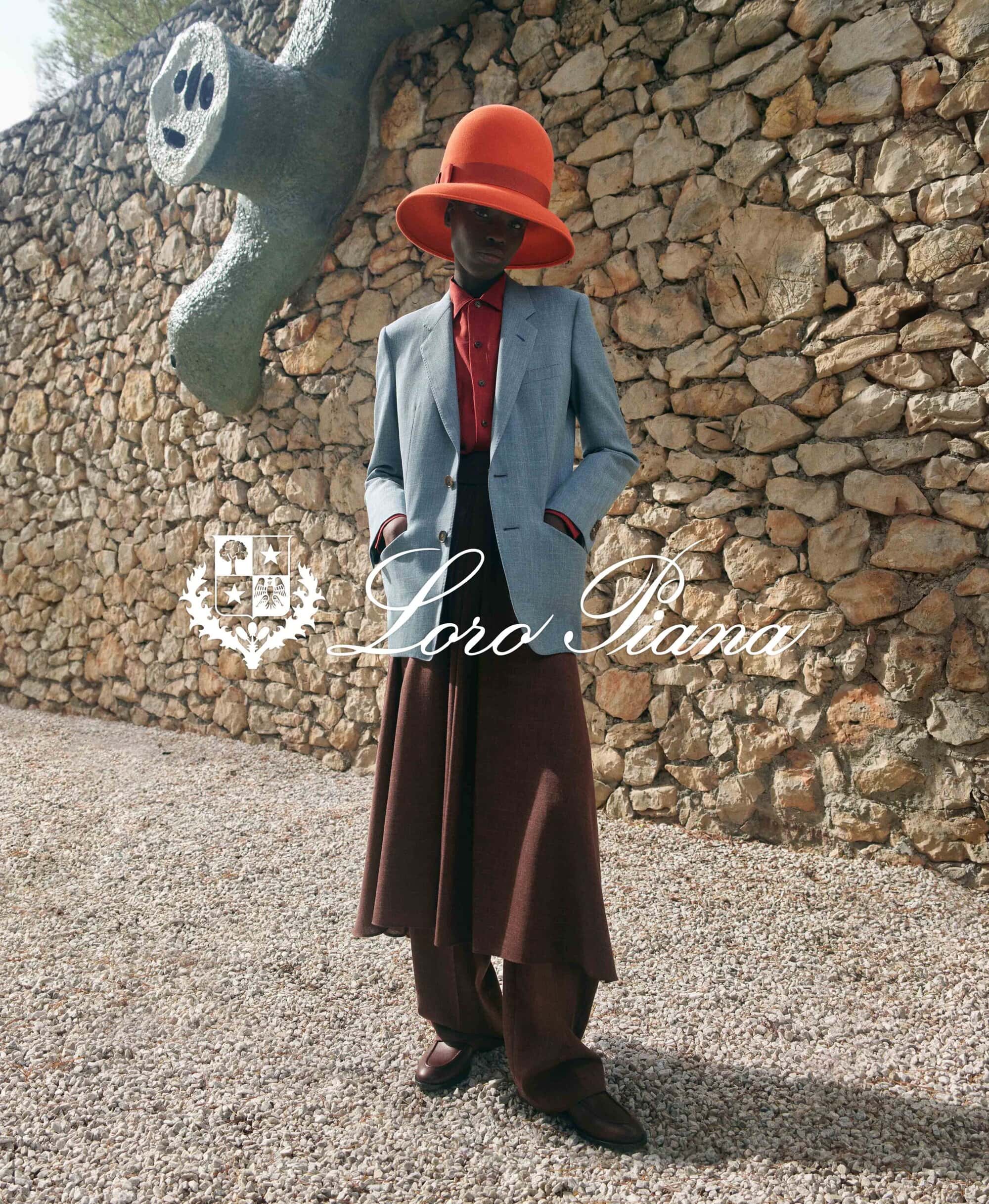 Stylish dark-skinned person in a blue blazer, red top, and large brown pants and hat poses in front of a stone wall with art.