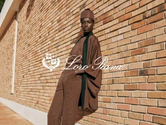 Model wearing a matching brown outfit and cap leaning against a textured brick wall for Loro Piana Spring 2026 Ad Campaign