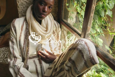 Model seated by a window holding a decorative object, wearing cream knit apparel with brown stripes for Loro Piana.