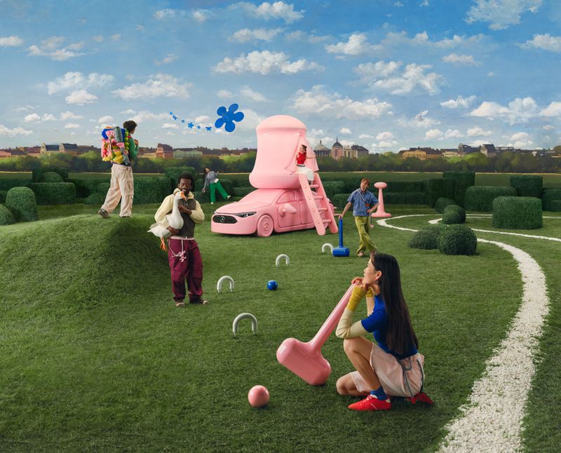 Group of people playing croquet with whimsical oversized mallets and a pink car-shaped playhouse under a blue sky with puffy clouds.