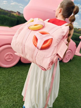 Woman carries a large pink quilted bag with colorful accents outside against a toy car and green fields under a cloudy sky.
