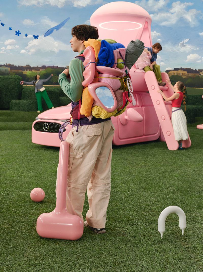 An individual with a unique backpack stands in a grassy field near a pink car and playground equipment, under a bright sky.