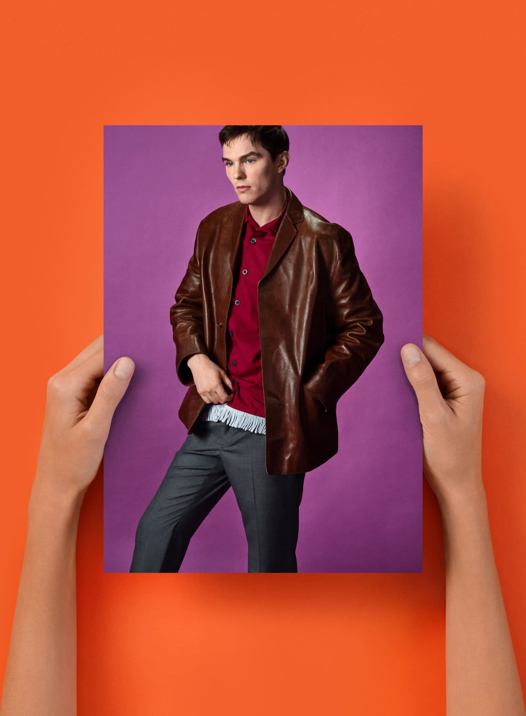 Stylish man poses in a studio-held photograph with a brown leather jacket, red shirt, and grey pants, set against a purple background.