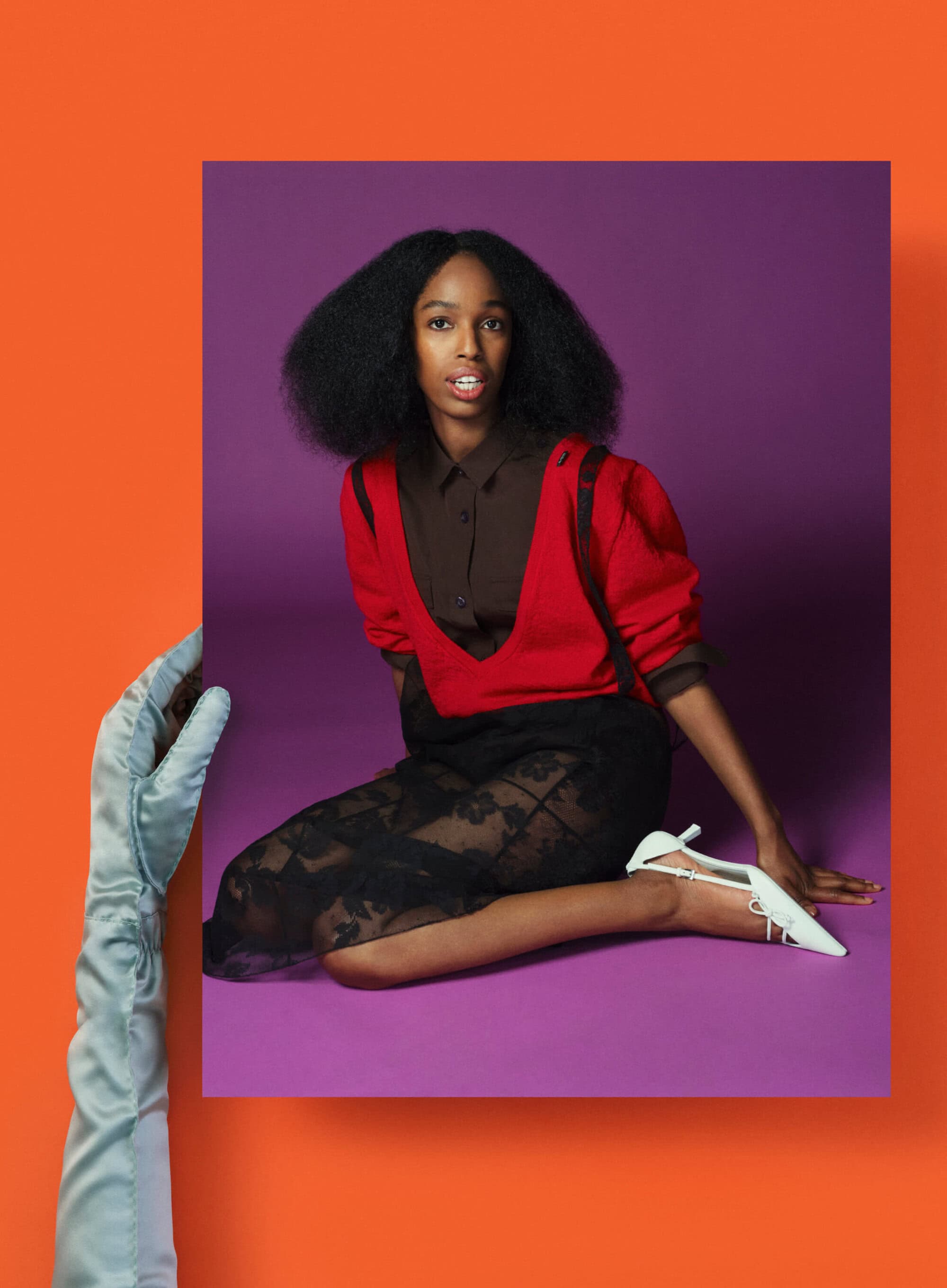 An elegant Black woman poses on a purple backdrop, wearing a red top and black lace skirt, in a fashion-forward portrait shot indoors.