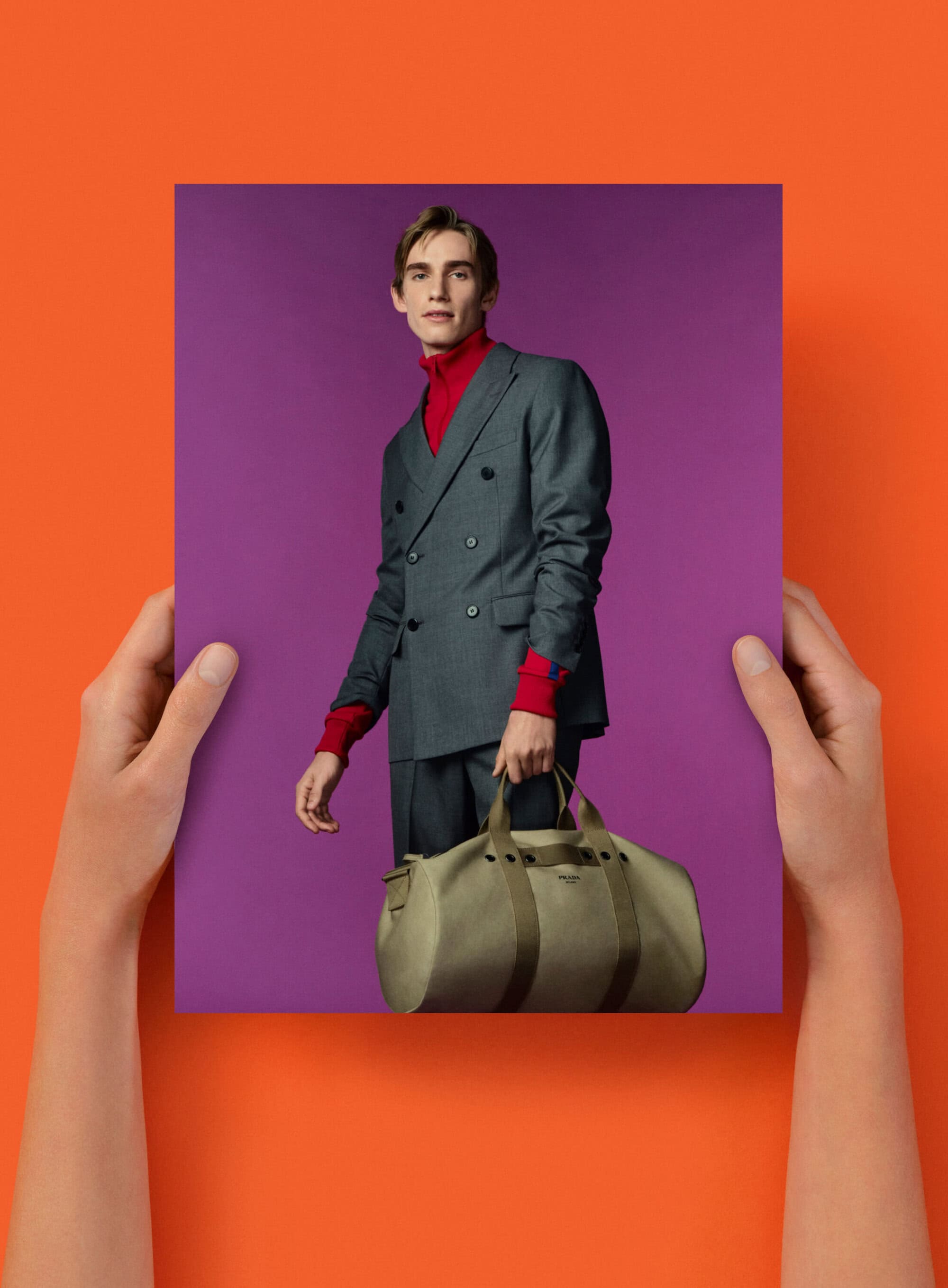 Fashion shot featuring a male model in a gray suit holding a duffel bag, presented on a purple backdrop against an orange background.