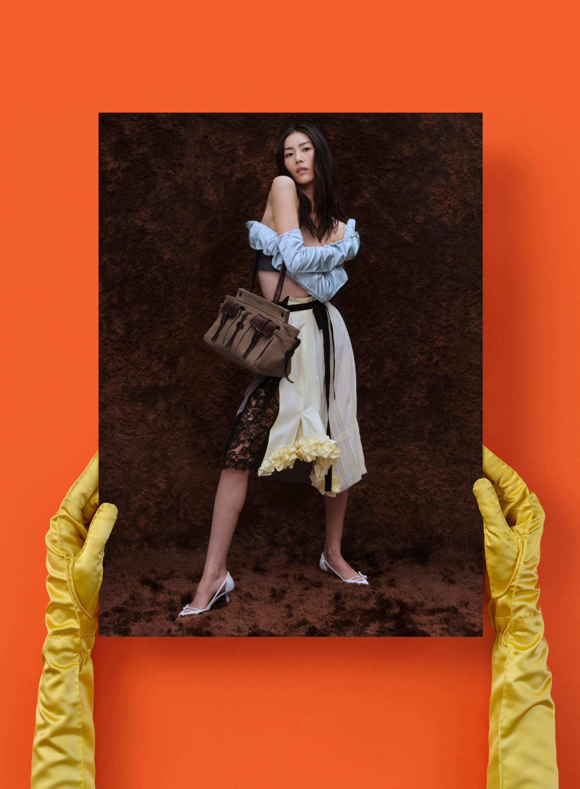 Woman posing with handbag in a framed photo held by yellow-gloved hands against an orange background, fashion portrait.